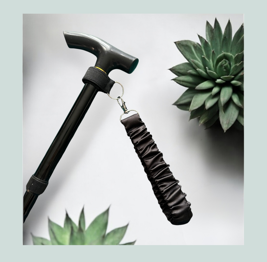 A close-up product shot of a black walking stick with a black satin wrist strap, attached with a black elastic wrap featuring hook and loop closure and metal  hardware, shown against a light gray background with decorative succulent plants.