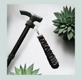 A close-up product shot of a black walking stick with a black satin wrist strap, attached with a black elastic wrap featuring hook and loop closure and metal  hardware, shown against a light gray background with decorative succulent plants.