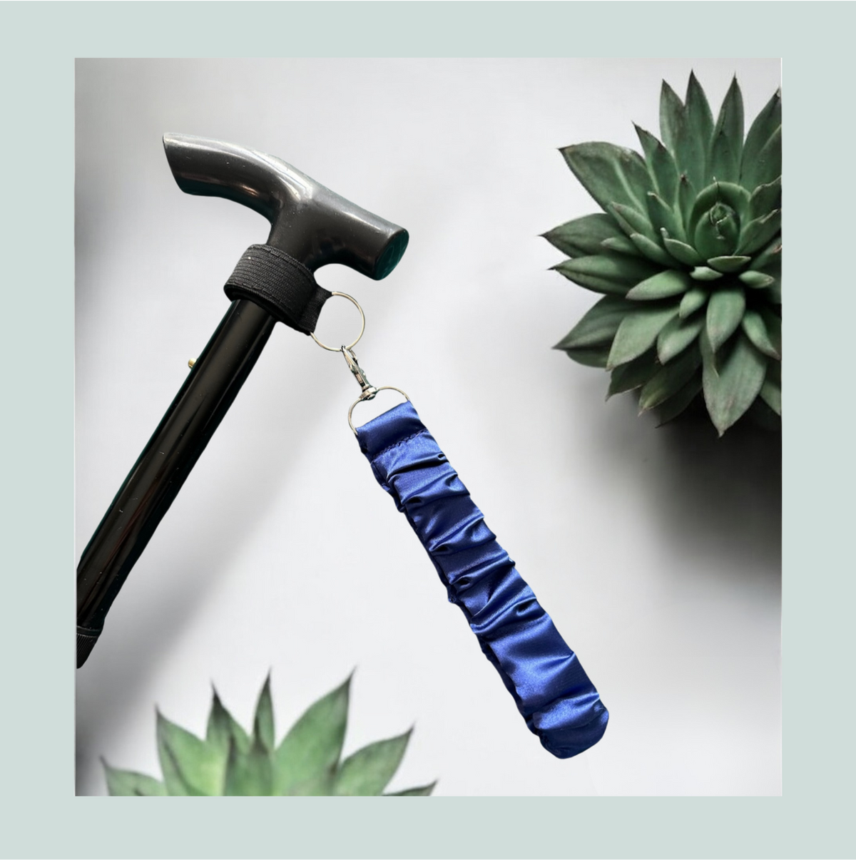 A close-up product shot of a black walking stick with a blue satin wrist strap, attached with a black elastic wrap featuring hook and loop closure and metal  hardware, shown against a light gray background with decorative succulent plants.