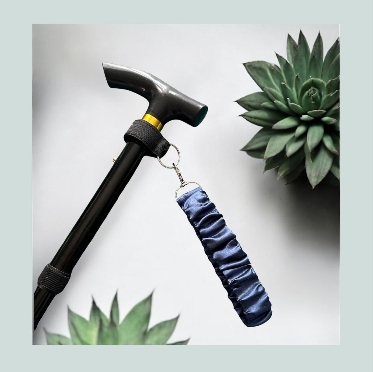 A close-up product shot of a black walking stick with a blue satin wrist strap, attached with a black elastic wrap featuring hook and loop closure and metal  hardware, shown against a light gray background with decorative succulent plants.