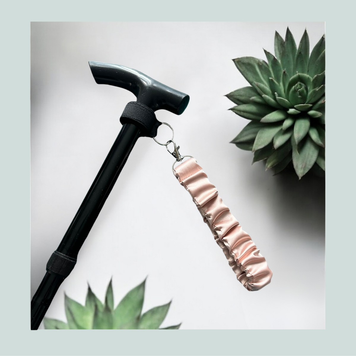 A close-up product shot of a black walking stick with a tea rose satin wrist strap, attached with a black elastic wrap featuring hook and loop closure and metal  hardware, shown against a light gray background with decorative succulent plants.