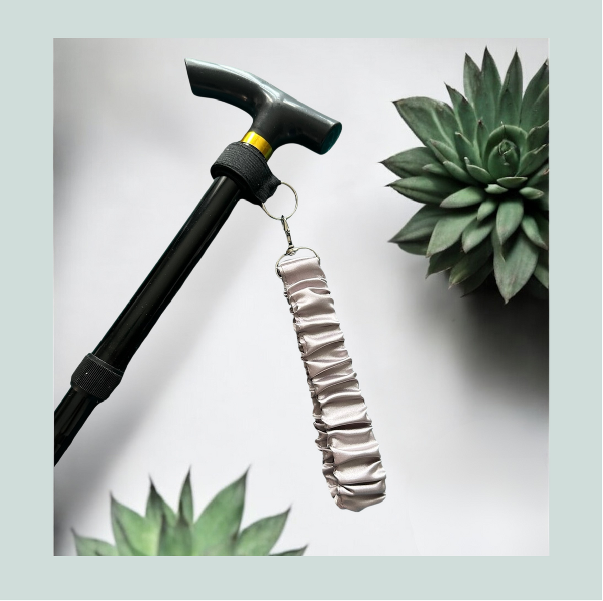 A close-up product shot of a black walking stick with a pale mauve satin wrist strap, attached with a black elastic wrap featuring hook and loop closure and metal  hardware, shown against a light gray background with decorative succulent plants.