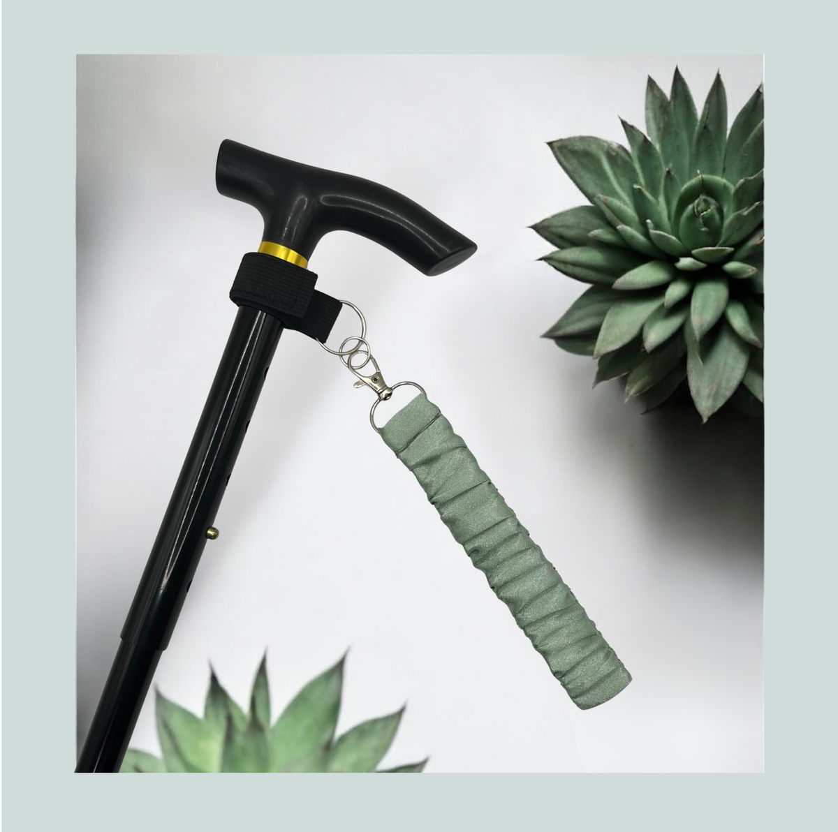 A black cane with a curved T-handle and a green fabric wrist strap, styled with succulent plants on a light gray background.