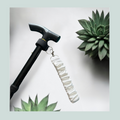 A close-up product shot of a black walking stick with a white satin wrist strap, attached with a black elastic wrap featuring hook and loop closure and metal  hardware, shown against a light gray background with decorative succulent plants.